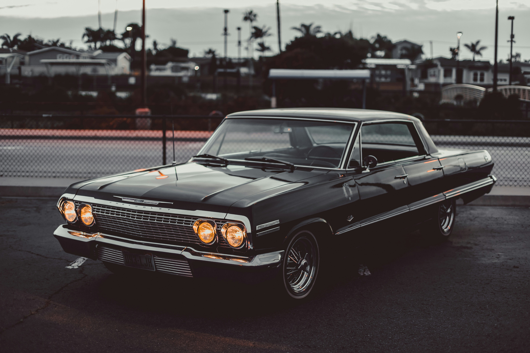 A classic black vintage car with its headlights on is parked on an asphalt surface near a fence, with palm trees and buildings visible in the background.