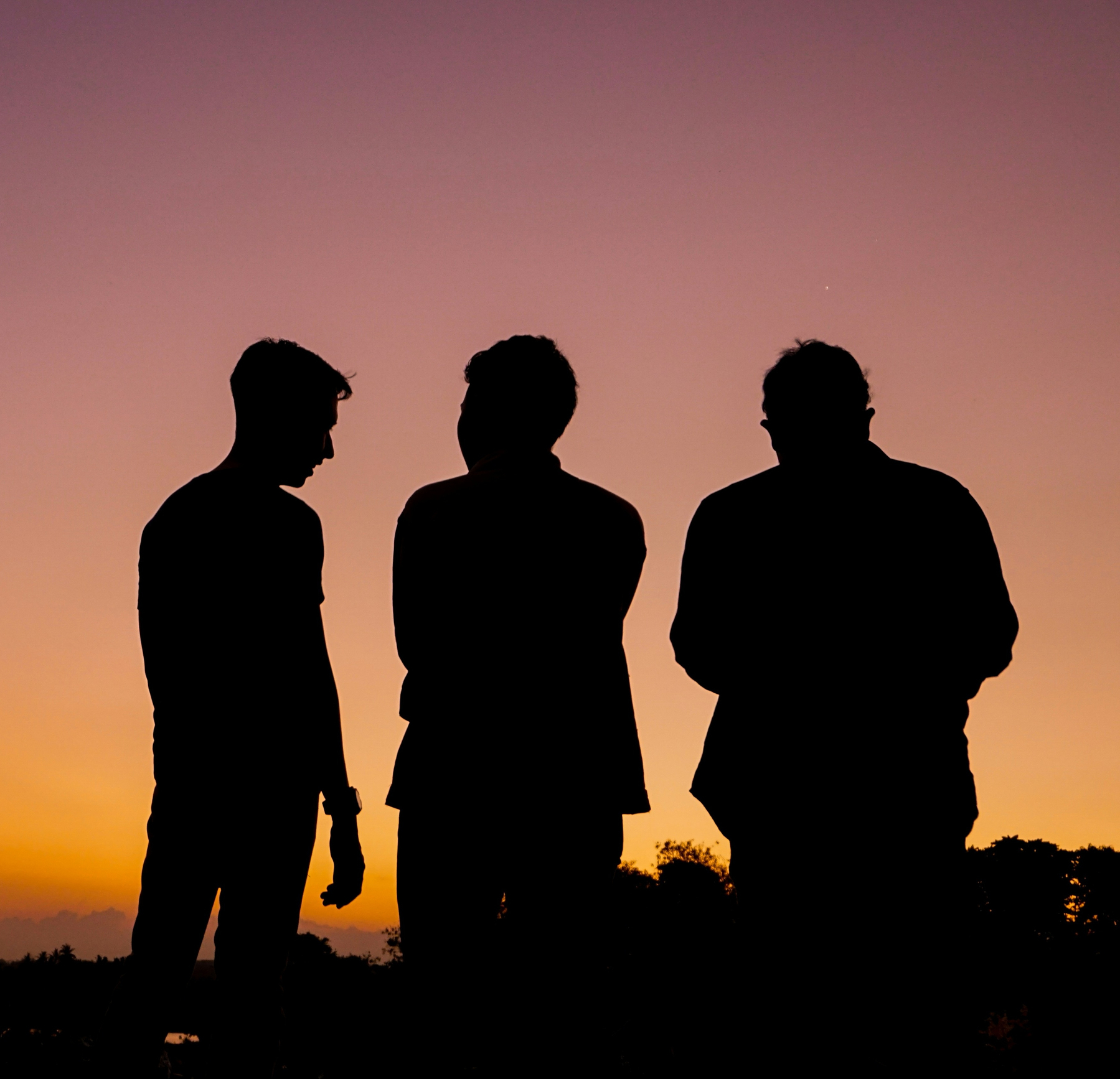 Three people are standing and facing each other against the backdrop of a colorful sunset sky.